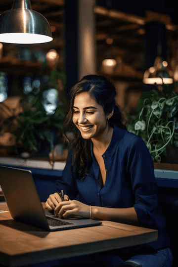 A cheerful woman in a blue shirt is typing on a laptop at a table in a cosy café environment, with plants all around and a hanging lamp overhead.